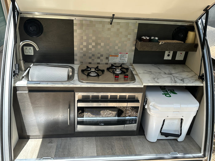 and a chair invite relaxation.|A compact RV rental teardrop trailer parked in a serene forest setting