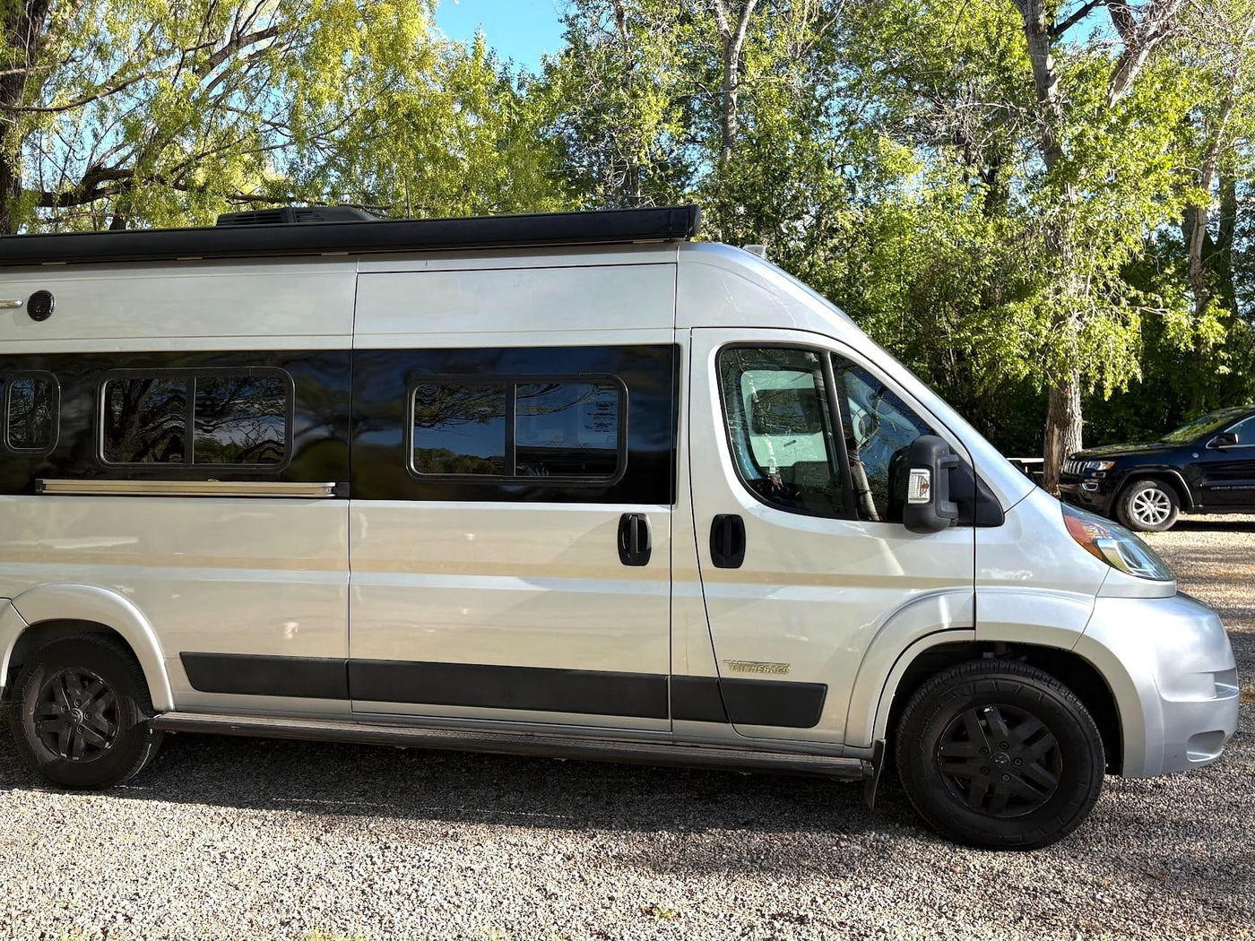Silver Winnebago Travato camper van, an ideal RV rental, parked on gravel amidst green trees. — photo 5