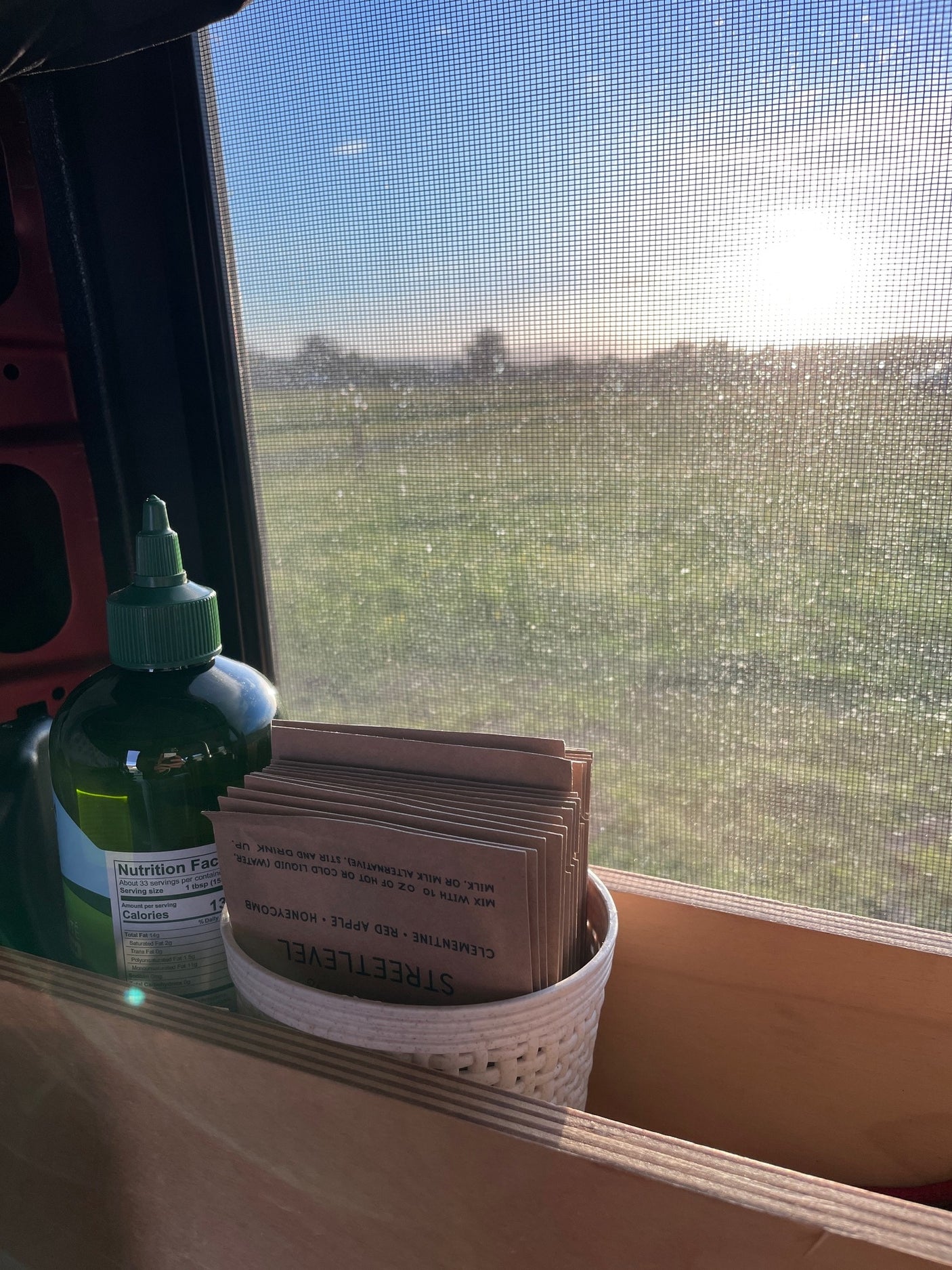 RV rental morning view: Sun through a screen, with a green bottle and 'Streetlevel' drink packets on a wooden shelf.