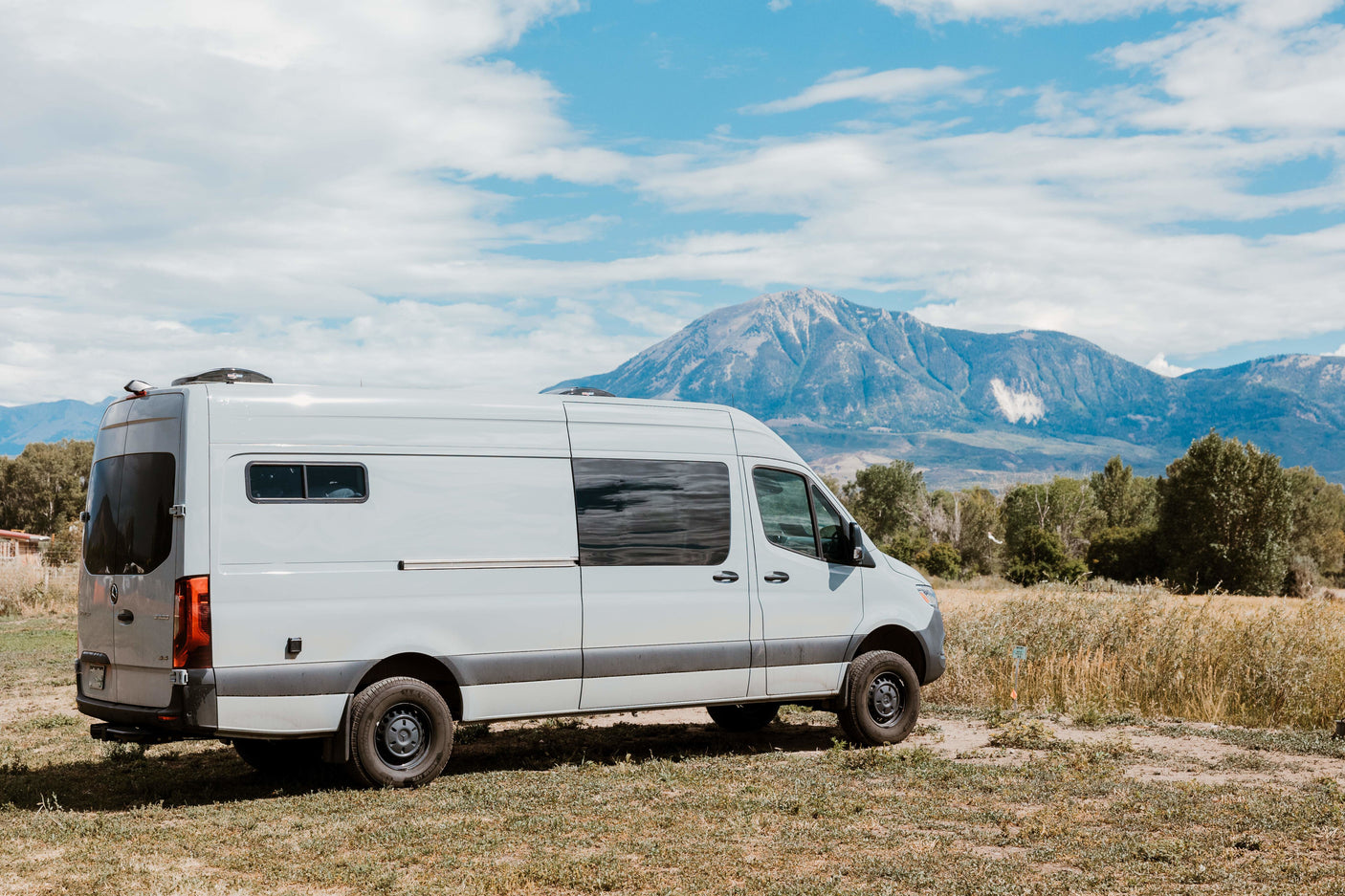 White RV rental van parked in a field with a majestic mountain and cloudy blue sky backdrop. Ideal for adventure travel.