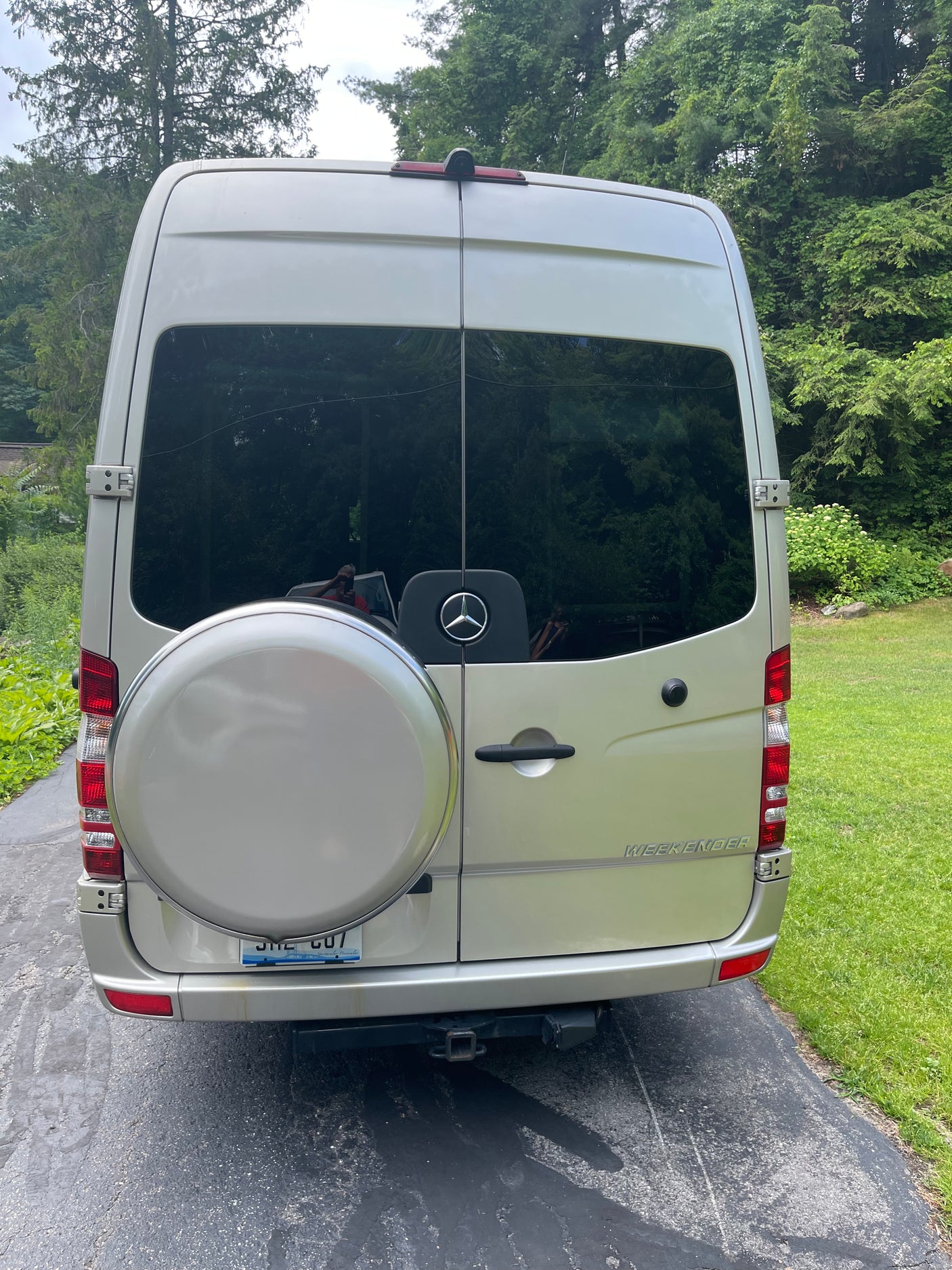 Rear view of a silver Mercedes-Benz Sprinter RV rental, with a spare tire & "WEEKENDER" badge, parked on an asphalt drive.