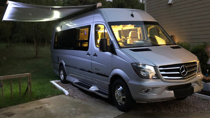 Silver Mercedes-Benz Sprinter RV rental parked at night, with an extended awning and illuminated interior.