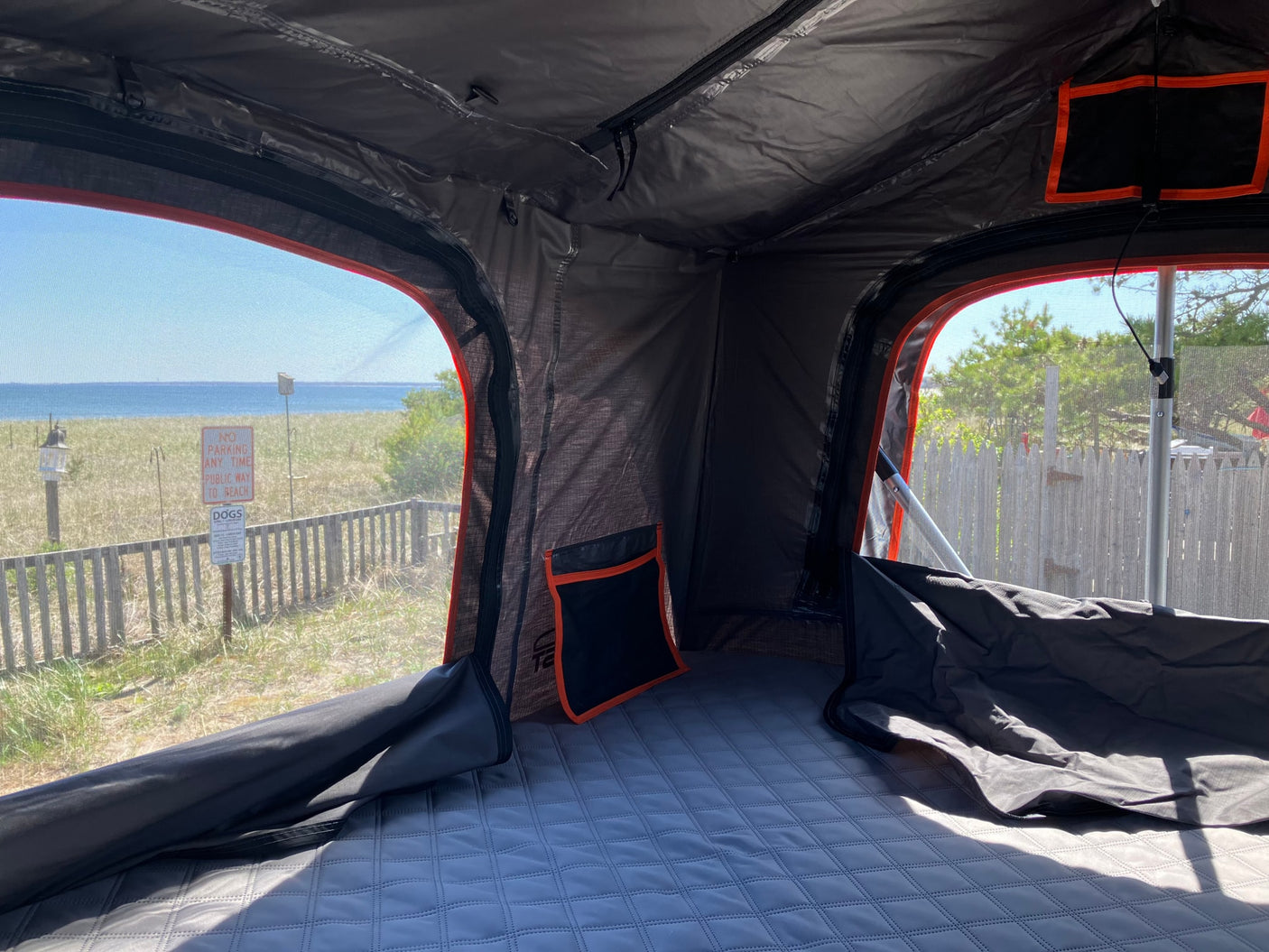 This comfortable sleeping space inside a rooftop tent, ideal for an RV rental trip, overlooks a sunny beach through its mesh windows.