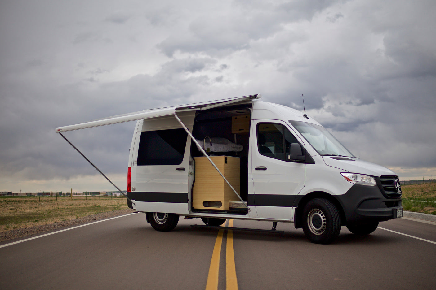 White Mercedes Sprinter RV rental with open side door, custom interior, and extended awning on a road. Ready for adventure!