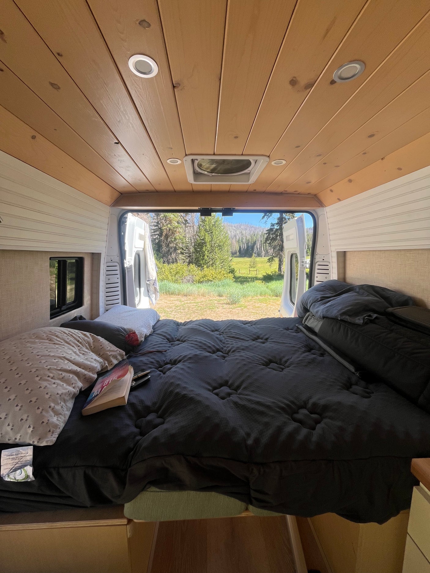 Cozy RV rental bed overlooks a serene forest and field through open rear doors. Perfect for a peaceful retreat.