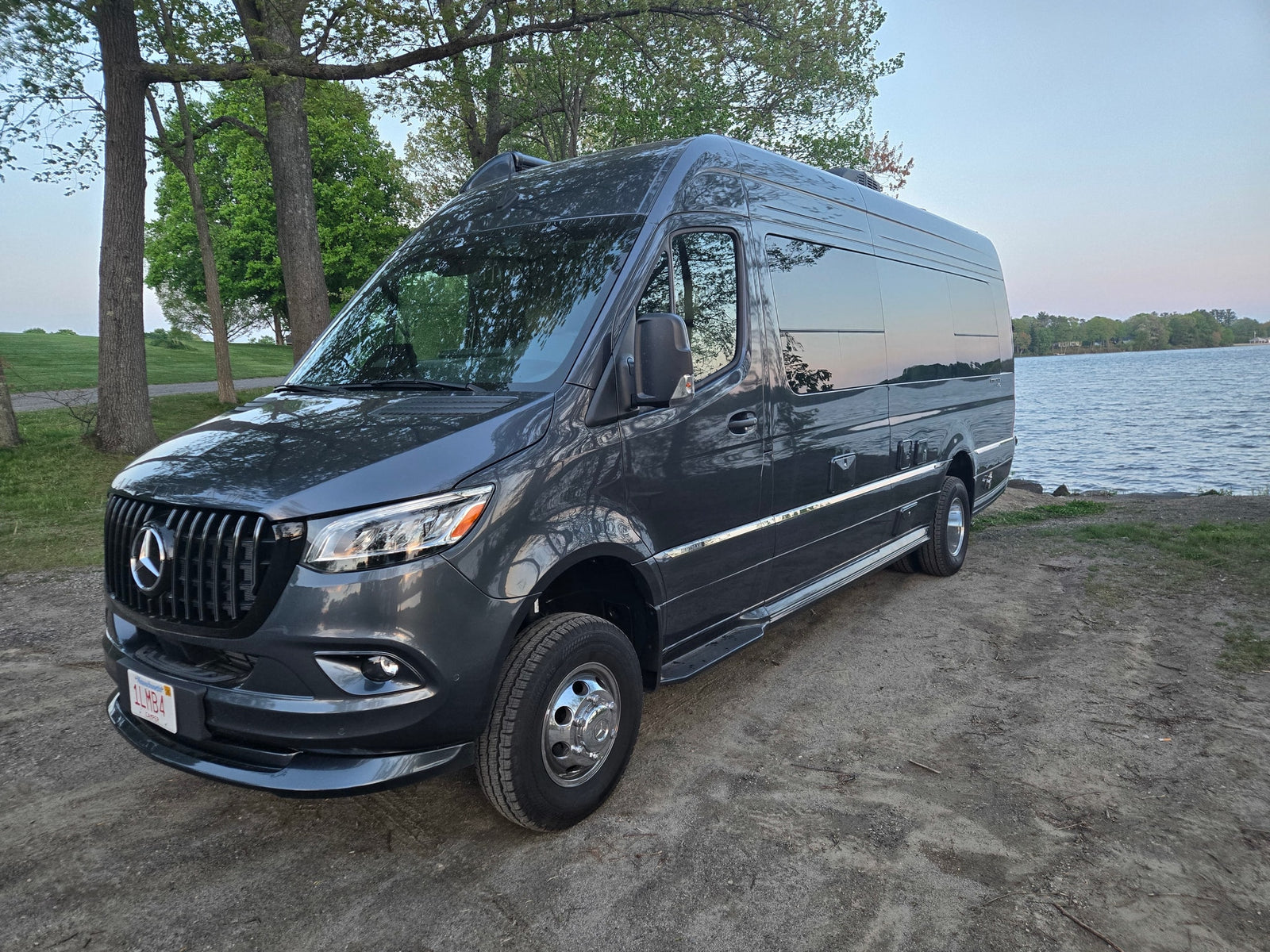 Dark gray Mercedes Sprinter rv rental camper van parked by a scenic lake under clear skies, surrounded by trees.