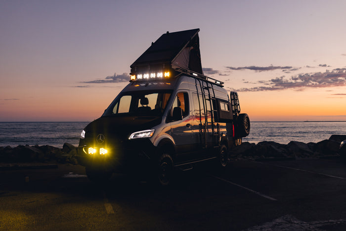 This adventure-ready Mercedes-Benz Sprinter RV rental, with a pop-top tent & bright lights, shines at sunset by the ocean.