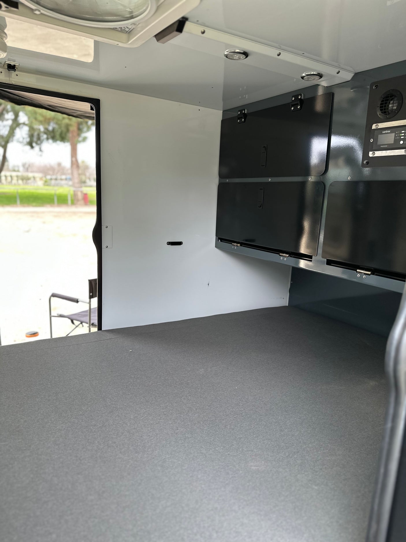 Inside an RV rental, featuring a grey sleeping platform, dark overhead storage, and a Xantrex electrical panel. Outdoor view.