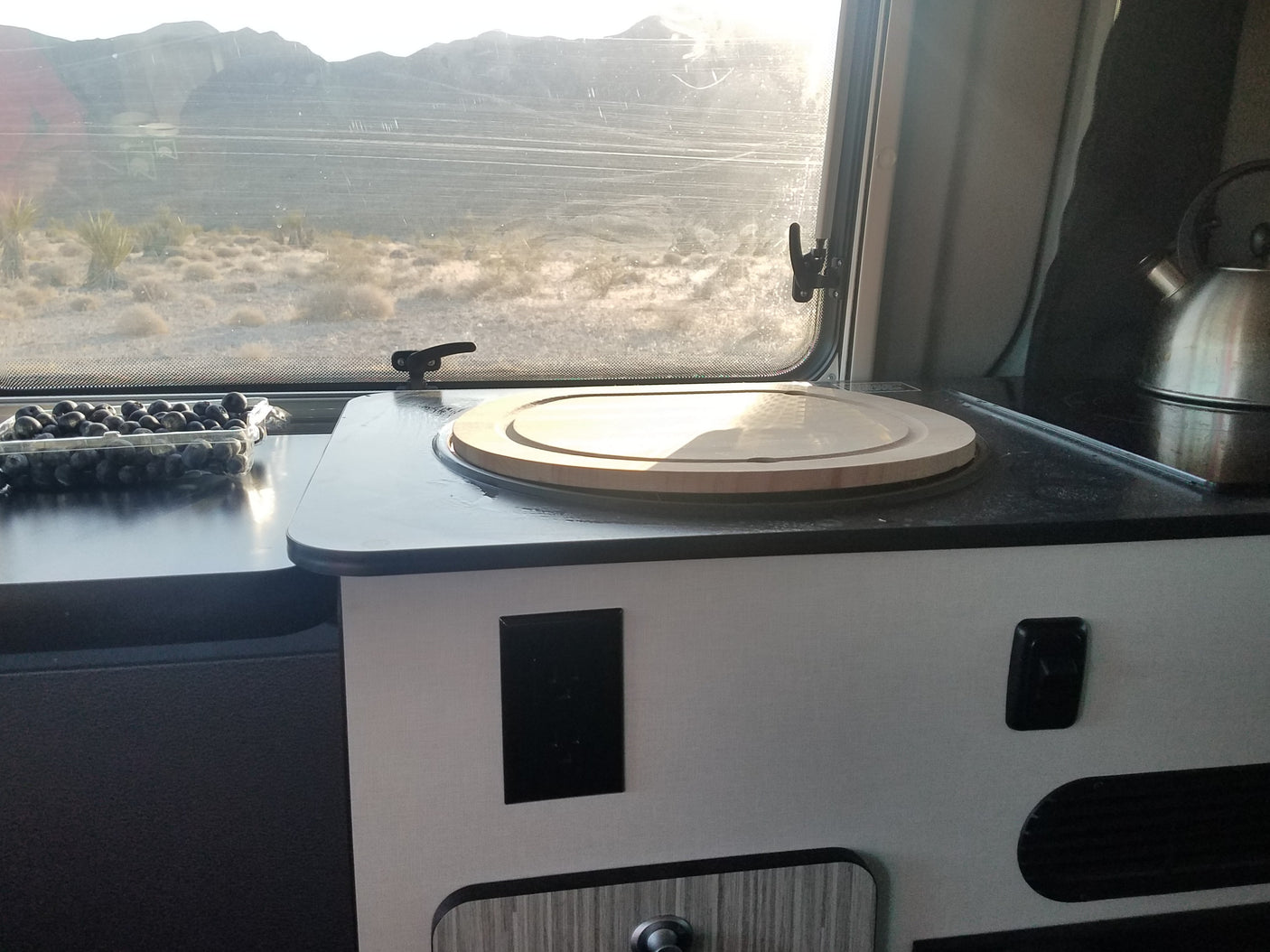 An RV rental's interior with counter, blueberries, and kettle, offering a bright desert landscape view from the window.