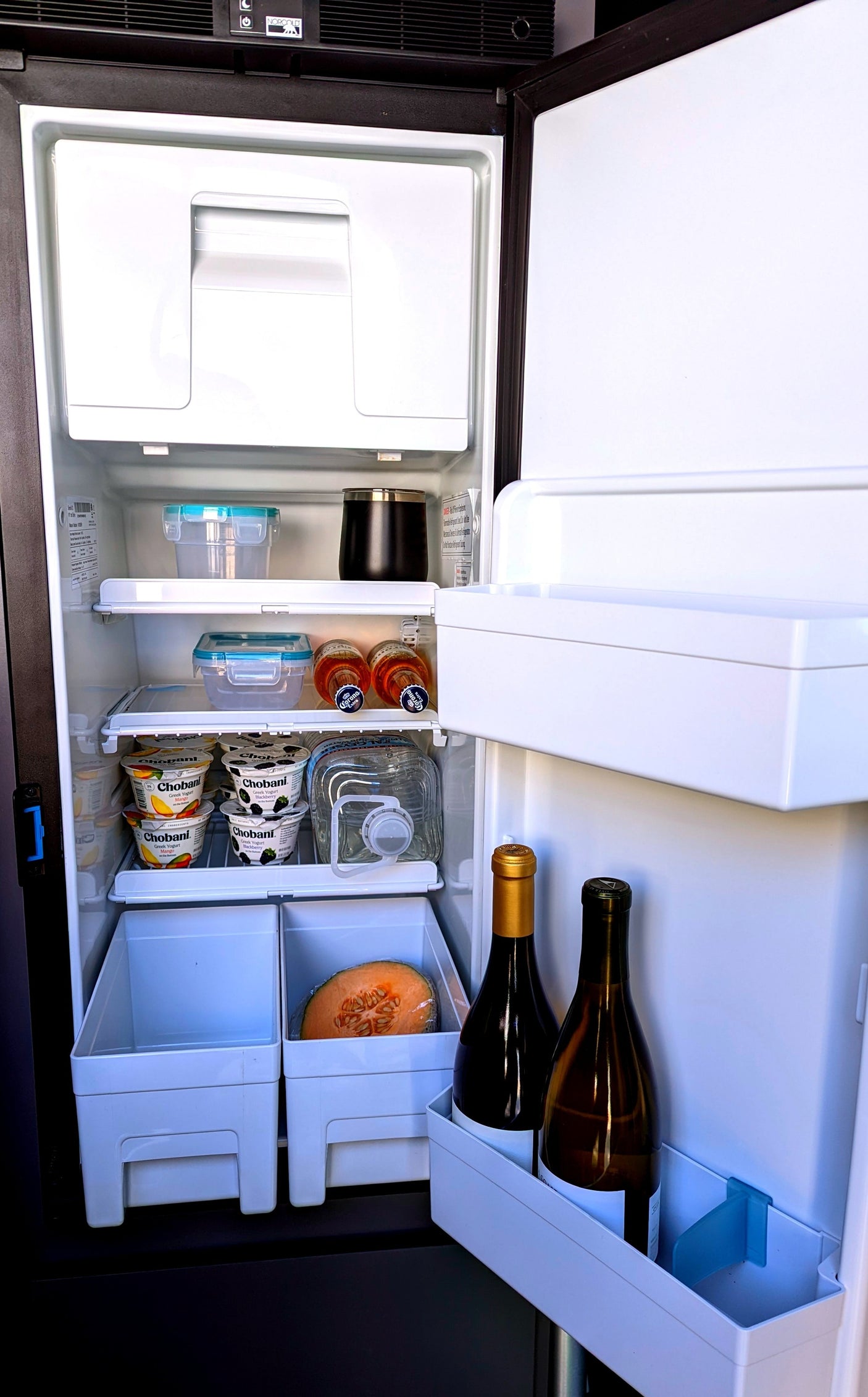 Inside an RV rental refrigerator, neatly organized shelves hold yogurts, drinks, and fruit, ready for road trip enjoyment.