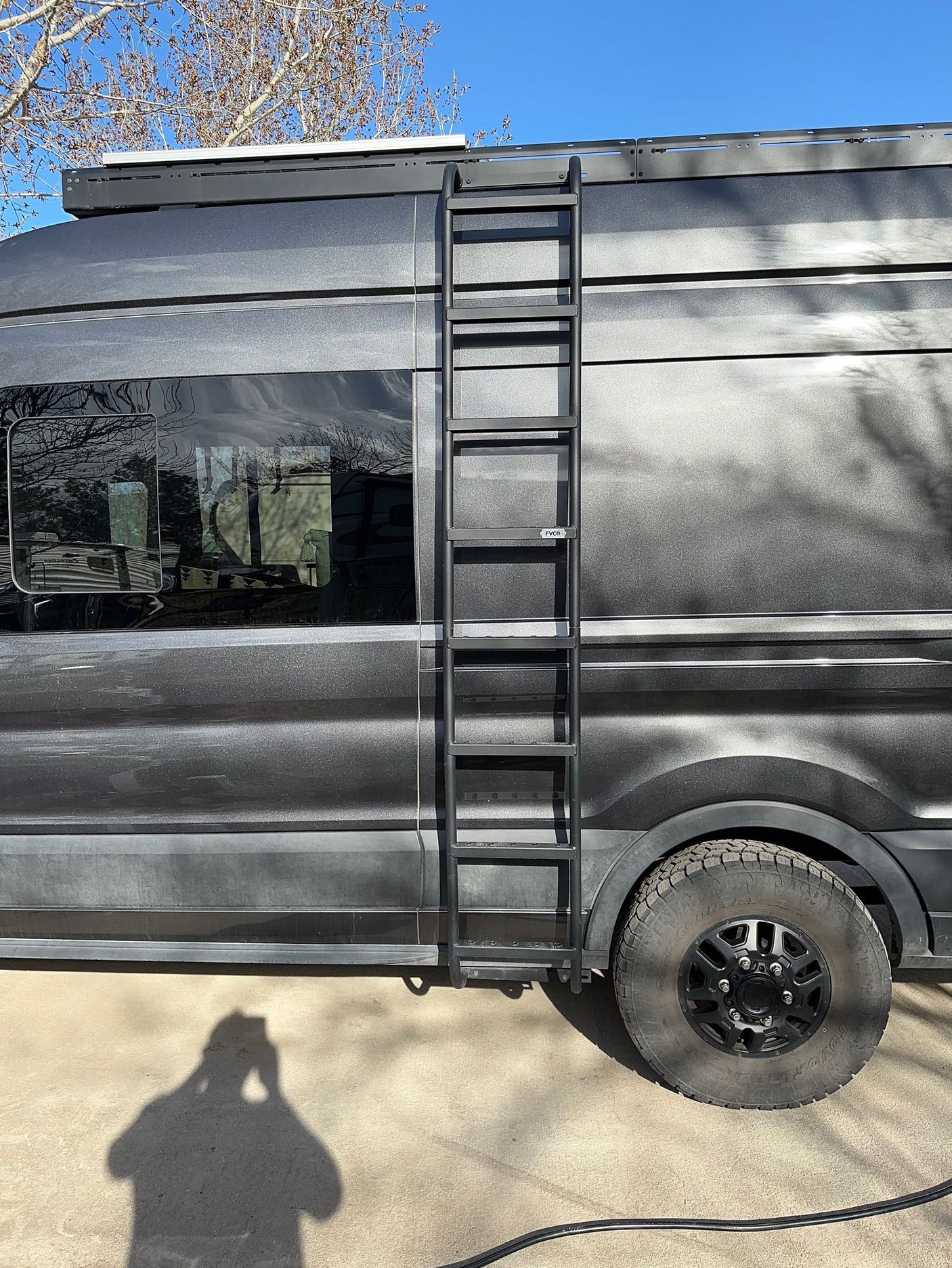 Close-up of a dark gray rv rental van, showing its side ladder, roof rack, large off-road tire, and textured body.