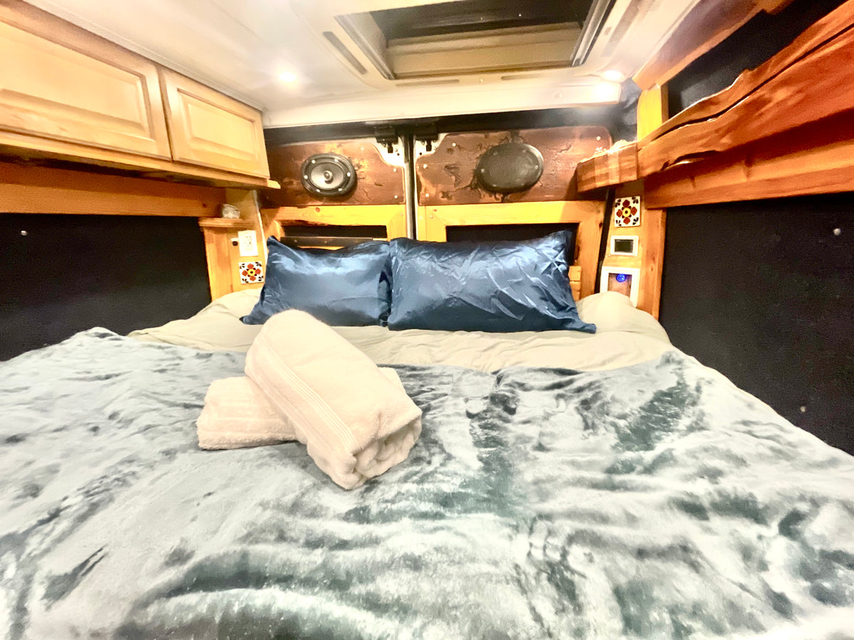 Cozy RV rental bed with blue satin pillows, fluffy blanket, and rolled towels. Wooden paneling and cabinets line the walls. — photo 18