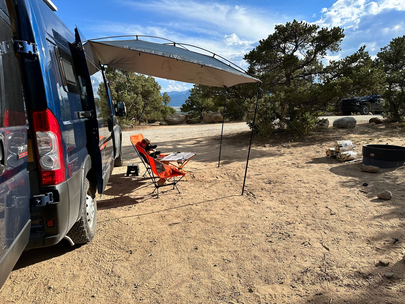 Relaxing at a dirt campsite with an RV rental. A blue camper van with an attached awning shades a person at a table.