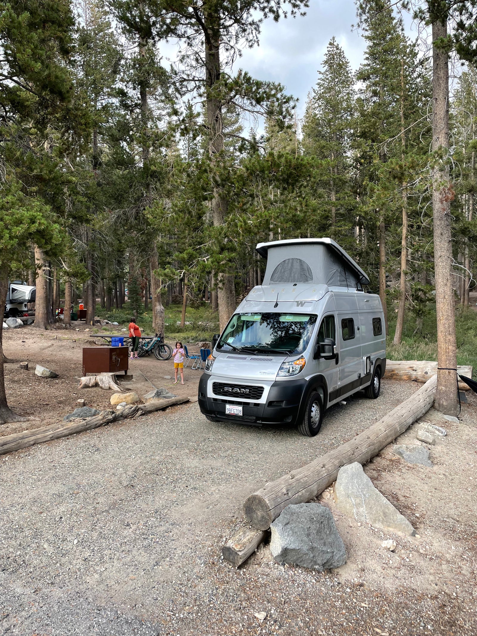 RV rental camping: Silver Winnebago camper van at a rustic site with people & bikes.
