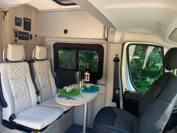 Inside an RV rental: a compact dining area with a table for two, plush seating, and controls. Enjoy nature through the windows.