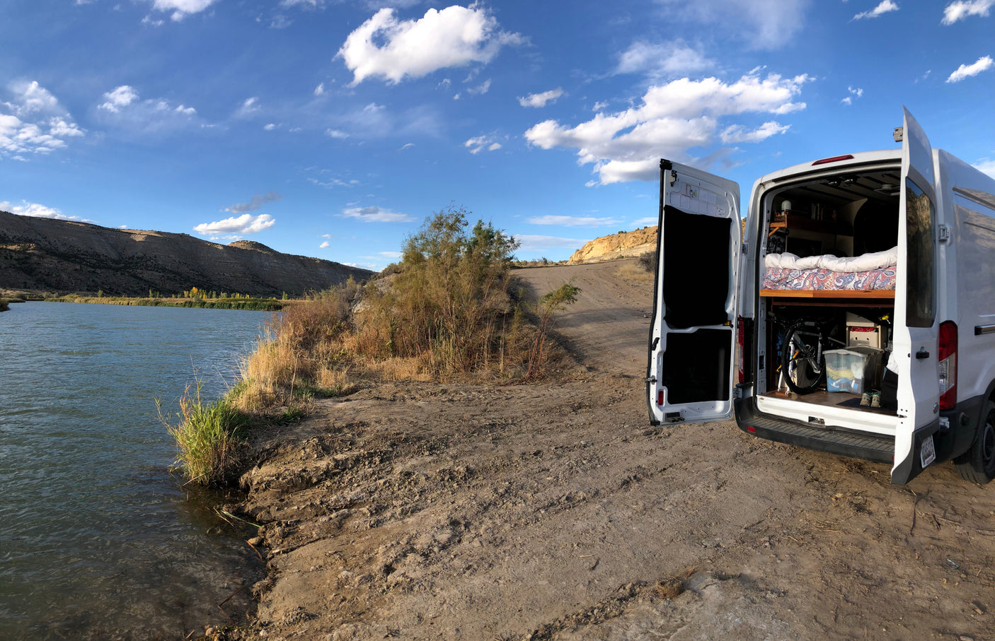 RV rental campervan at a scenic river spot, rear doors open revealing the cozy bed, bike & gear inside, ideal for camping.