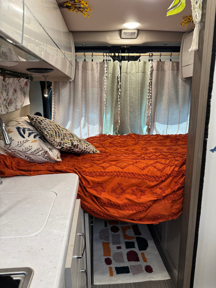 Cozy RV rental interior: orange bed with pillows, sunlit curtains. Counter & geometric rug visible, creating a homey feel.