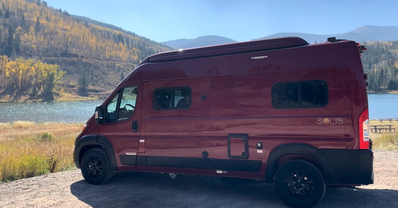 Maroon Winnebago Solis campervan by a fall lake. Explore nature with an RV rental.