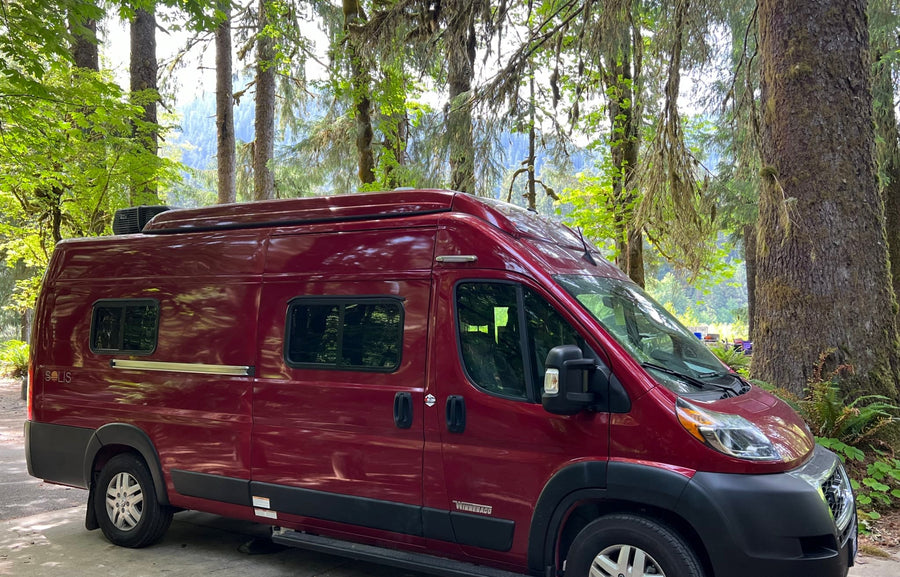 A red Winnebago Solis RV rental is parked in a serene forest campground, ideal for nature adventures.
