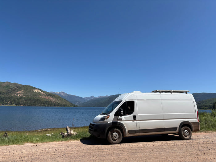 White RV rental van by a blue lake & green mountains under a clear sky. A dog peeks from the window, enjoying the scenic view.