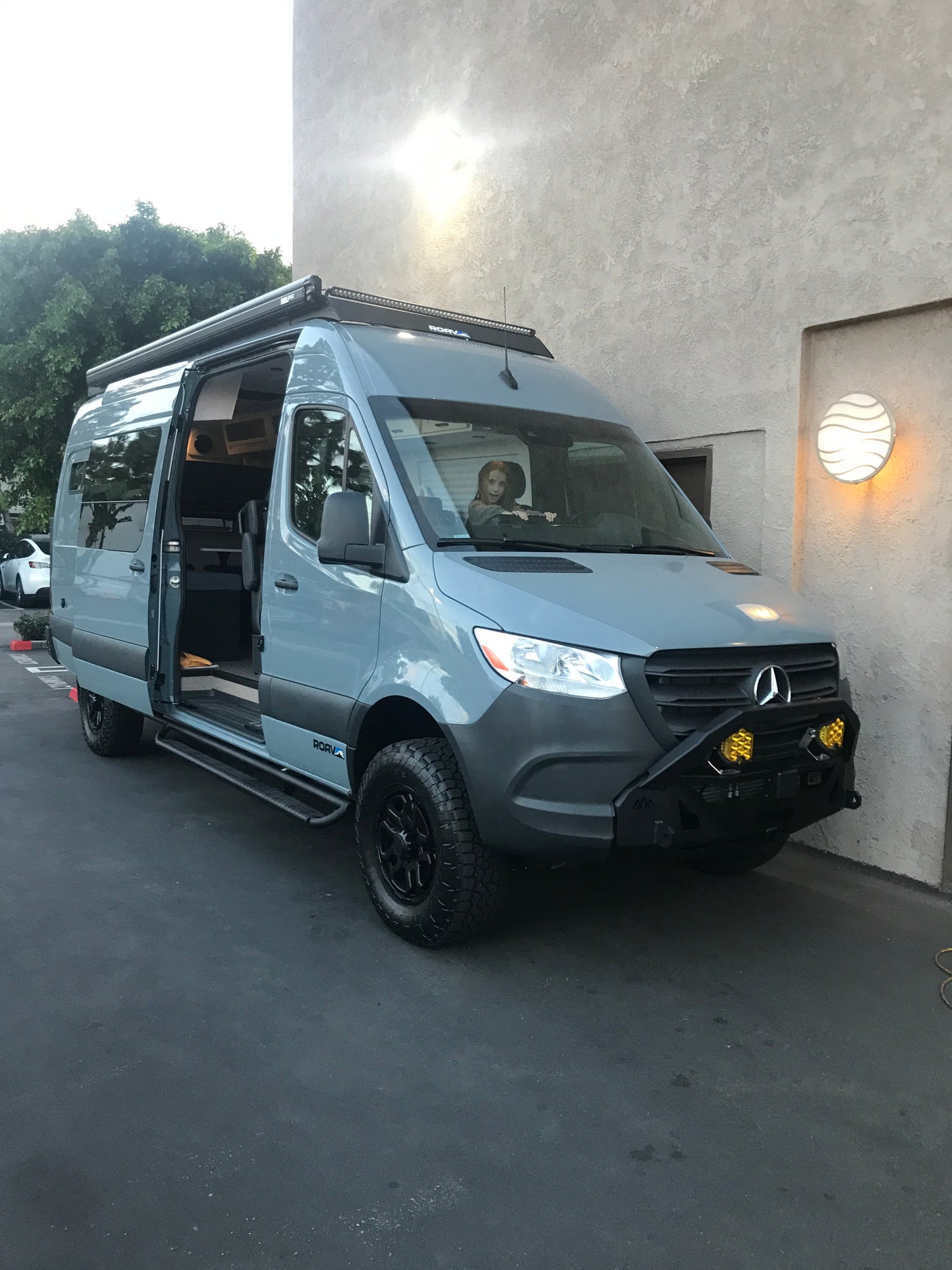 Custom grey Mercedes Sprinter RV rental. Child in driver's seat. Features off-road tires, light bar, and open side door.