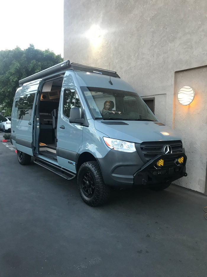 Custom grey Mercedes Sprinter RV rental. Child in driver's seat. Features off-road tires, light bar, and open side door.