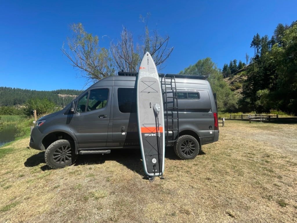 Experience van life! Gray camper van and paddleboard by water.