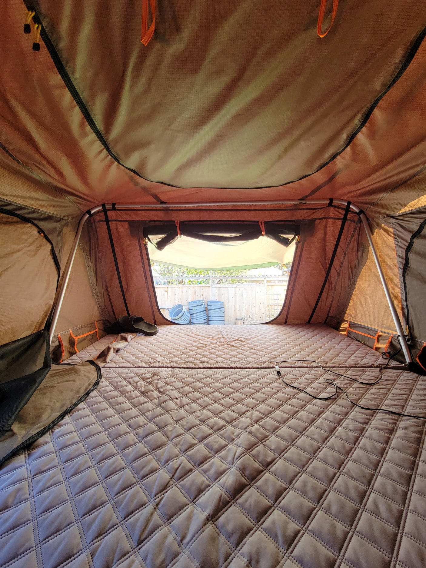 View inside a cozy RV rental rooftop tent: a quilted mattress on the floor and windows showing a backyard fence.