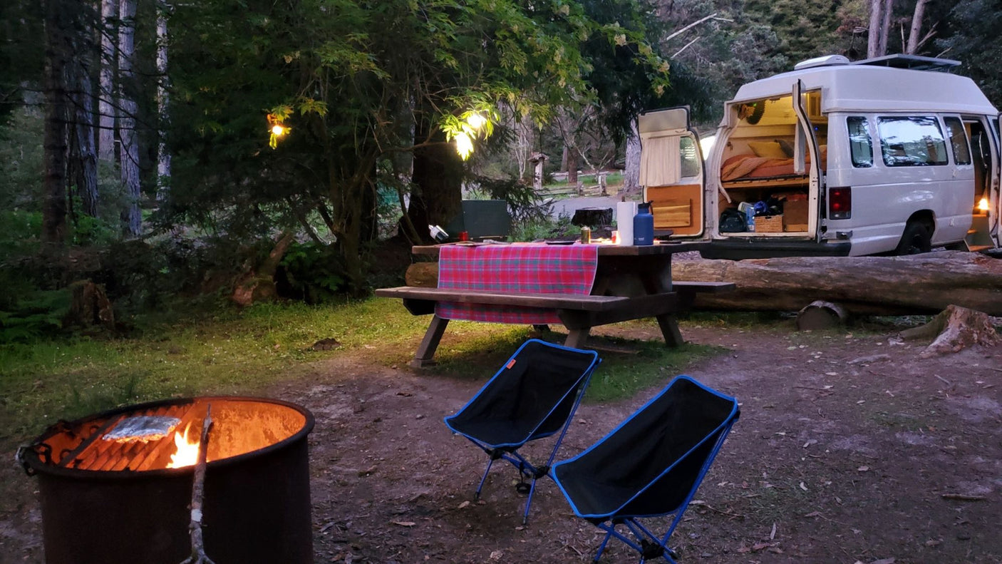 RV rental campsite featuring a camper van, picnic table, chairs, and a warm campfire under string lights at dusk.