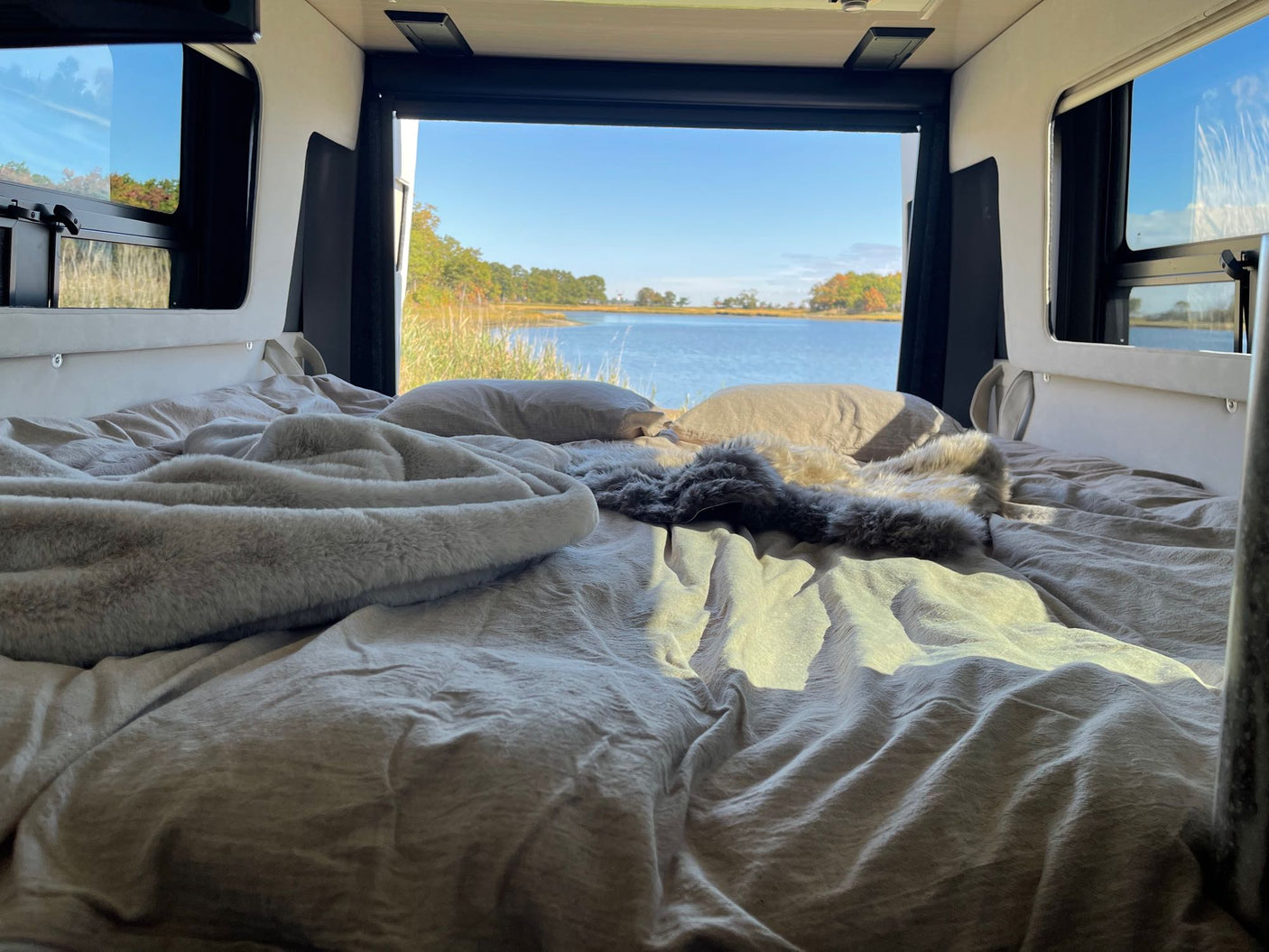 Cozy RV rental bed facing a stunning calm bay with trees. Soft blankets and pillows invite relaxation.