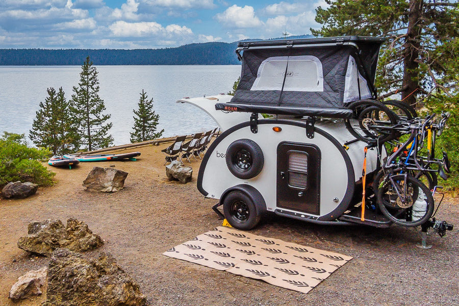 An RV rental teardrop trailer with a rooftop tent and bikes is parked at a scenic lake campsite with paddleboards.