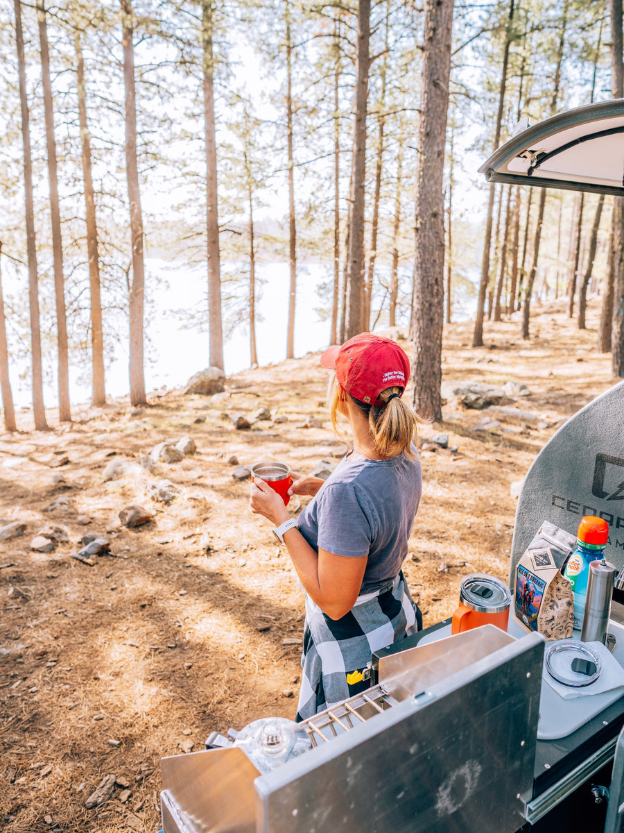 Woman sips coffee by a scenic lake, enjoying an RV rental camping trip among the pine trees.