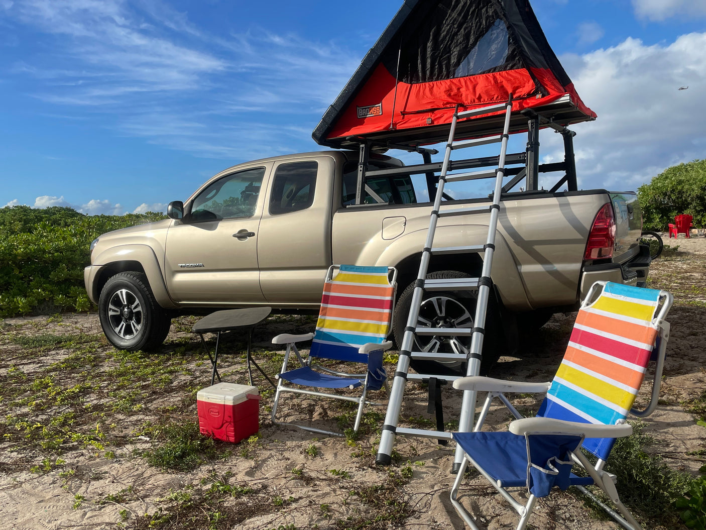 A tan pickup truck with a red rooftop tent and ladder, set up as an rv rental for camping. Beach chairs and cooler on sandy ground.