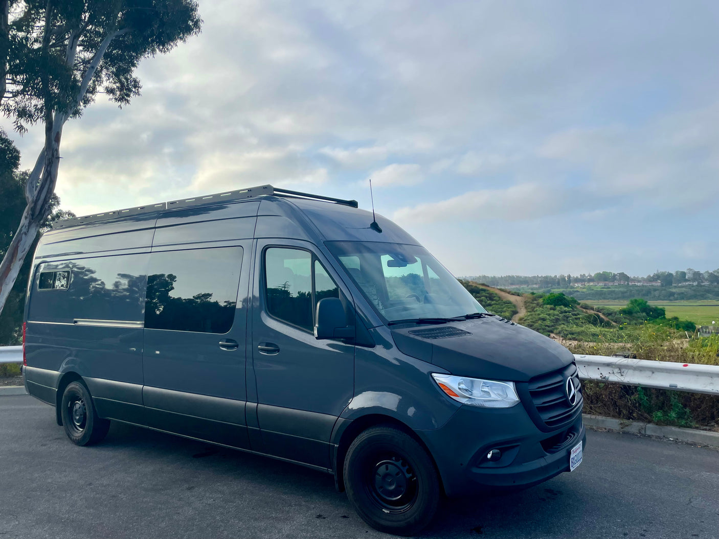 Dark gray Mercedes-Benz Sprinter camper van, ideal for rv rental, parked on a scenic road overlooking green hills under cloudy skies.
