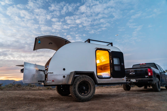 An RV rental teardrop camper, with its illuminated sleeping area and open rear kitchen, hitched to a pickup truck for off-grid camping.