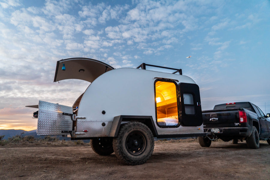 An RV rental teardrop camper, with its illuminated sleeping area and open rear kitchen, hitched to a pickup truck for off-grid camping.