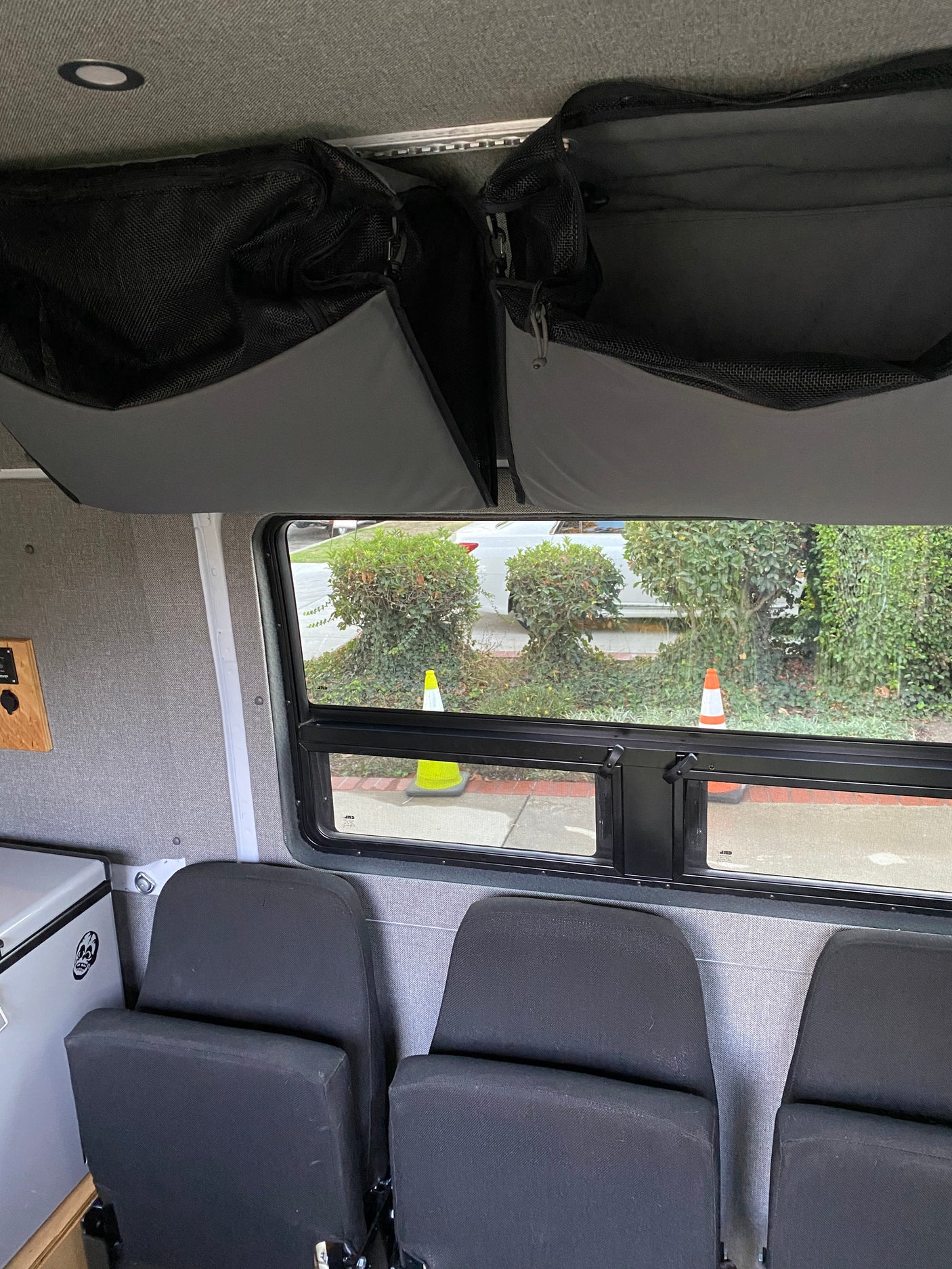RV rental: Overhead mesh storage, window to outdoor traffic cones/bushes, passenger seats, and cooler visible.