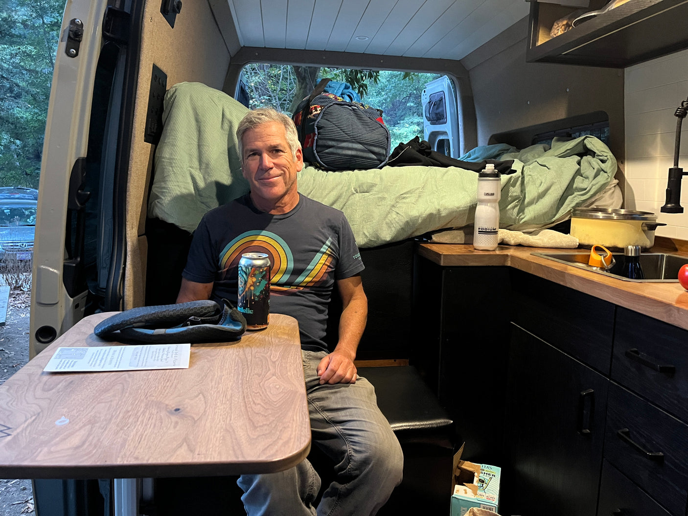 Man sips a drink in a cozy camper van rental, featuring a smart interior with a bed, table, and kitchen area.