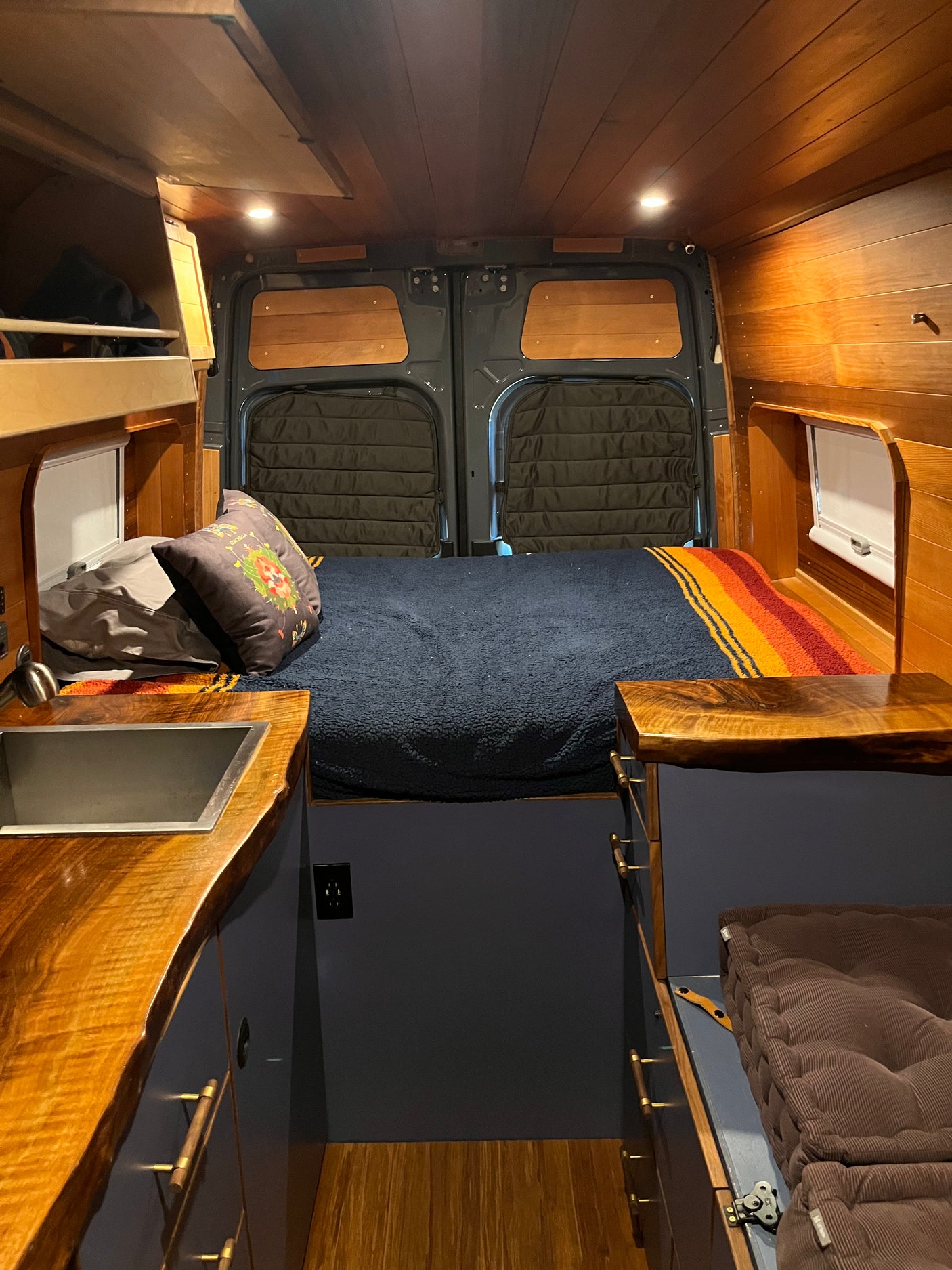 Cozy RV rental interior: wood-paneled walls, comfortable bed, custom storage, and rear doors in a camper van.