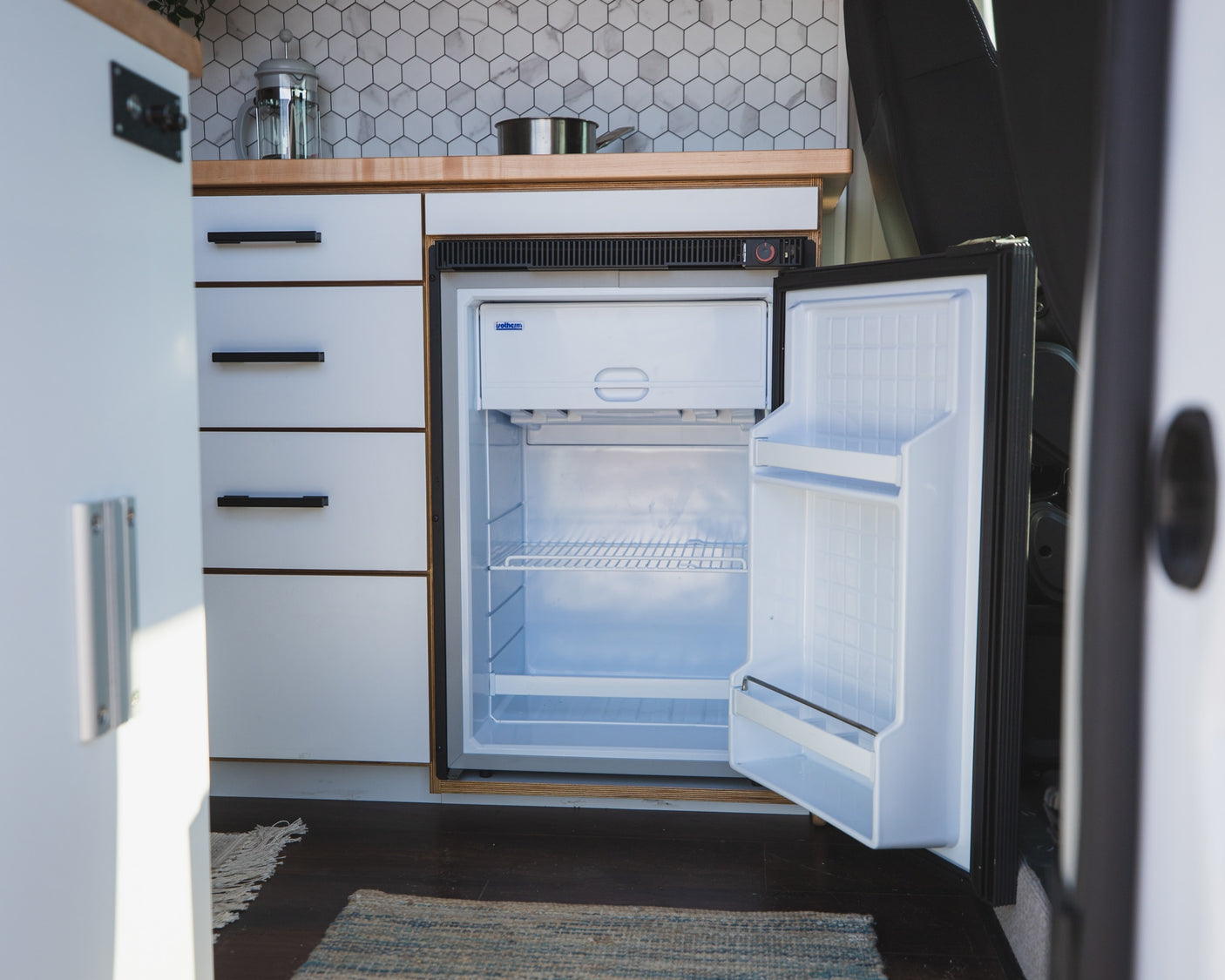 RV rental kitchen showcasing a compact, open refrigerator, white cabinets, wood counter, and tile backsplash.