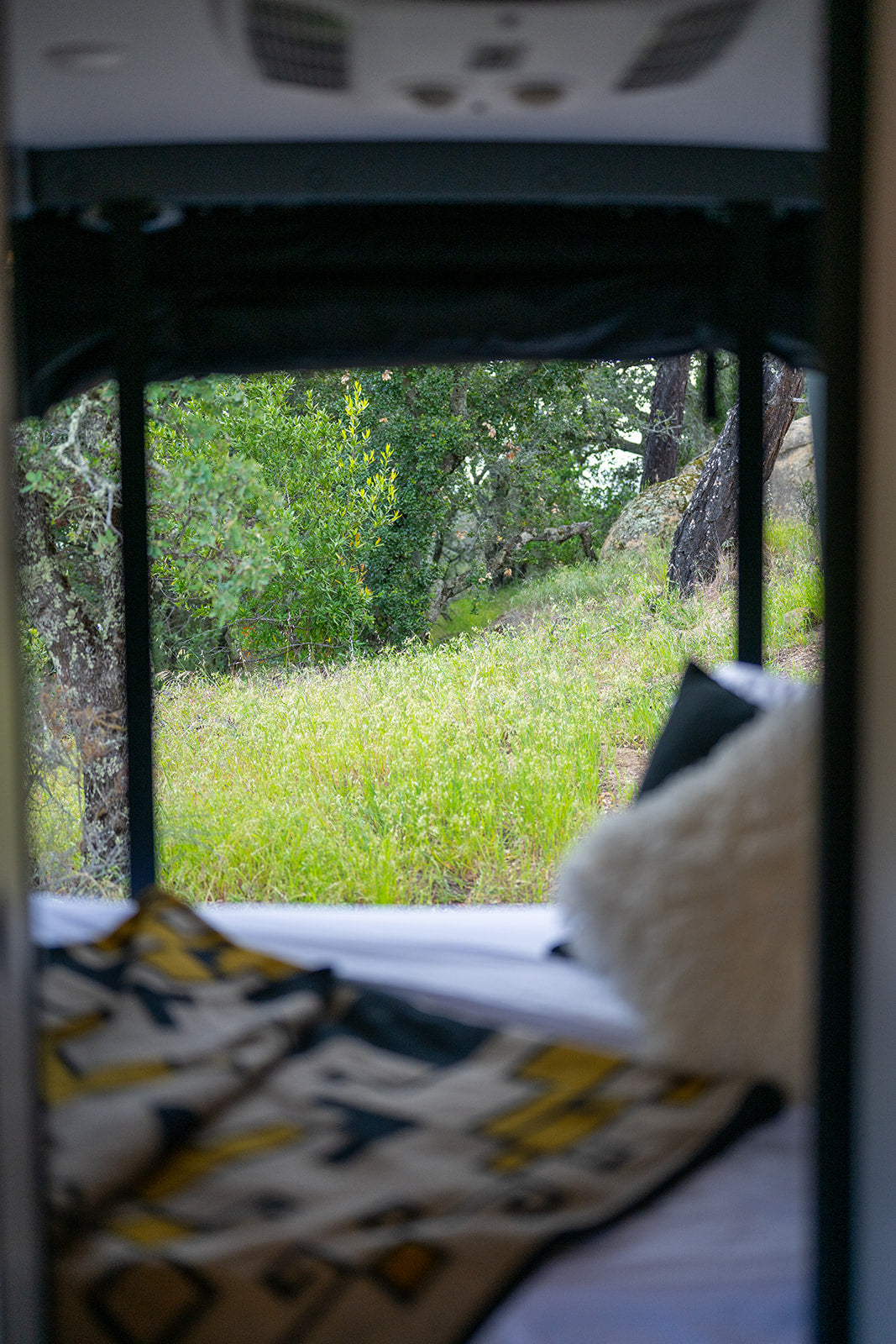 A view from inside an RV rental bed reveals vibrant green nature: trees, grass, and rocks outside the window.