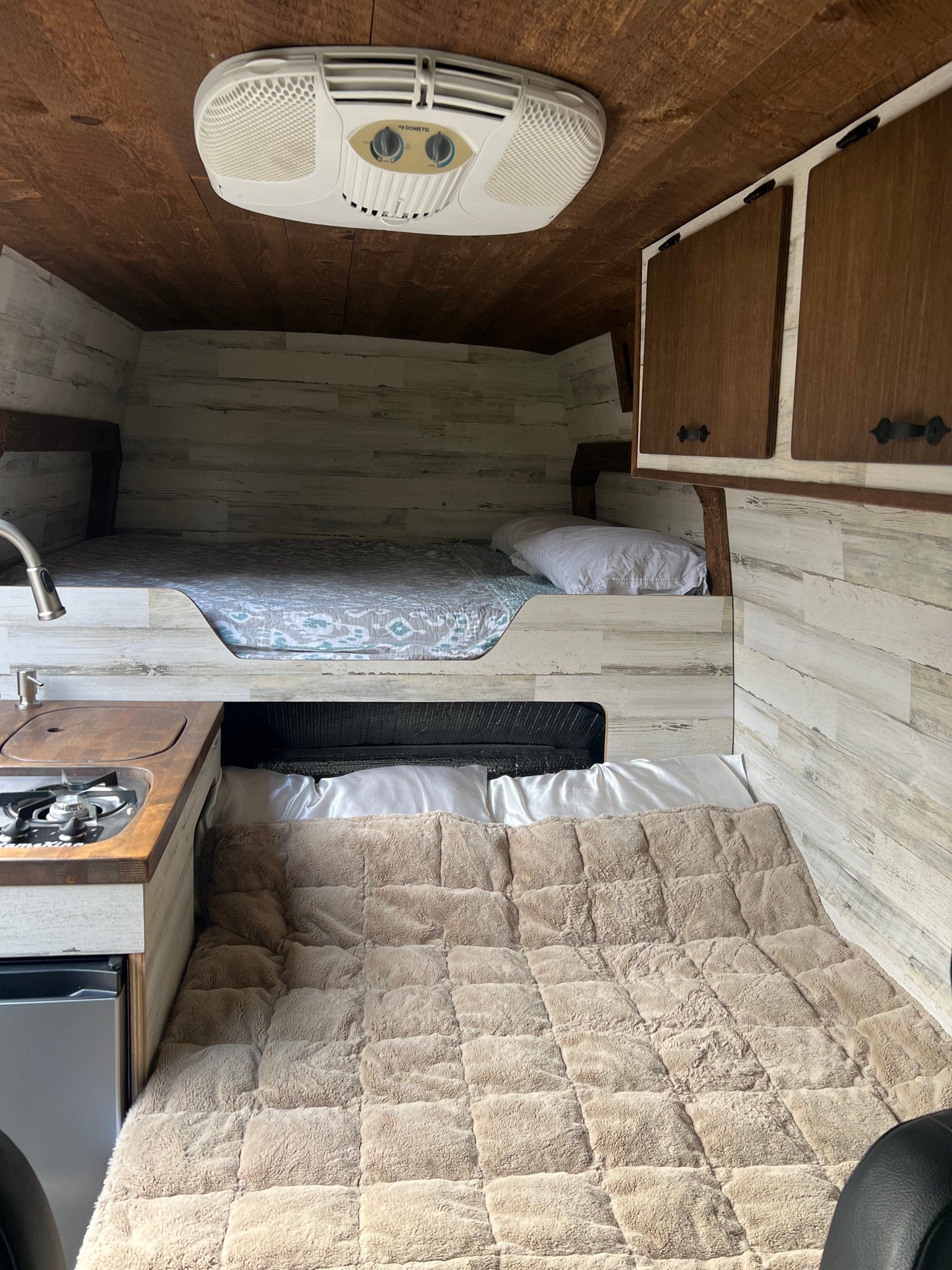 Inside an RV rental: two comfy bunk beds, compact kitchen with fridge, and AC unit. Walls feature distressed white wood paneling.