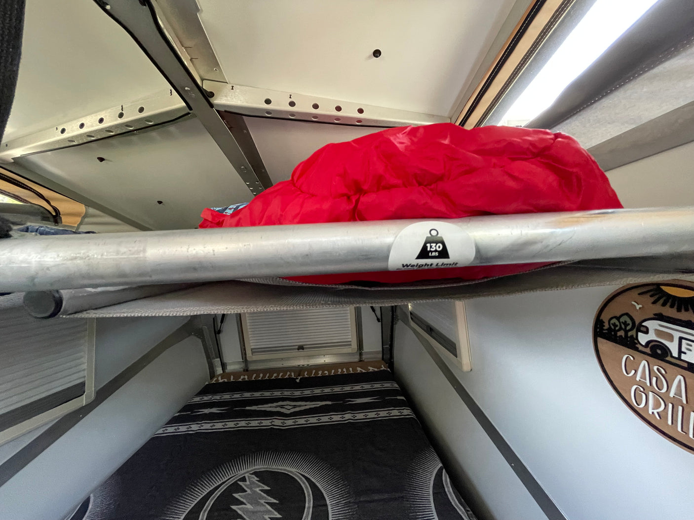 Inside an RV rental: a top bunk with a 130 lbs weight limit and red sleeping bag, over a bed with a patterned blanket.
