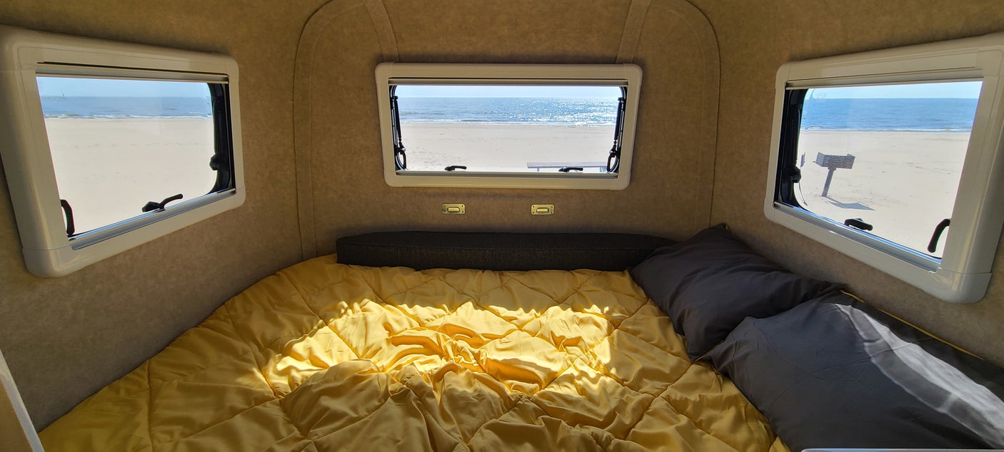 Beachfront RV rental: Wake up to ocean views from this cozy camper bedroom with a yellow bed and open windows overlooking a sandy shore.