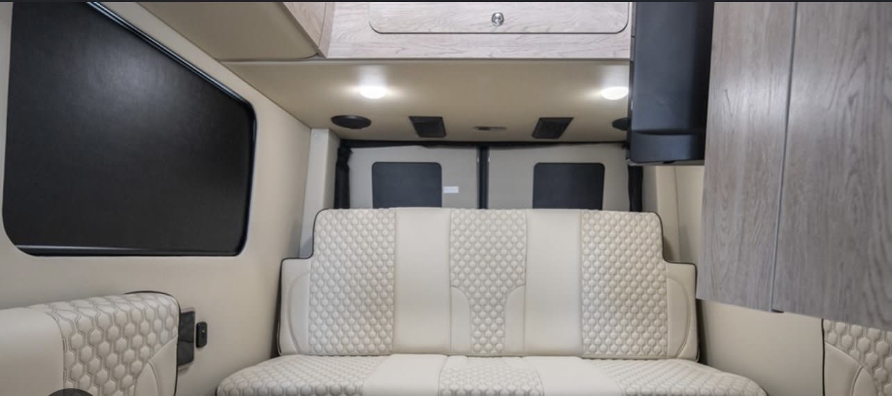 Modern rv rental interior: light-colored quilted seating, overhead cabinets, and dark window blinds. Ready for travel.