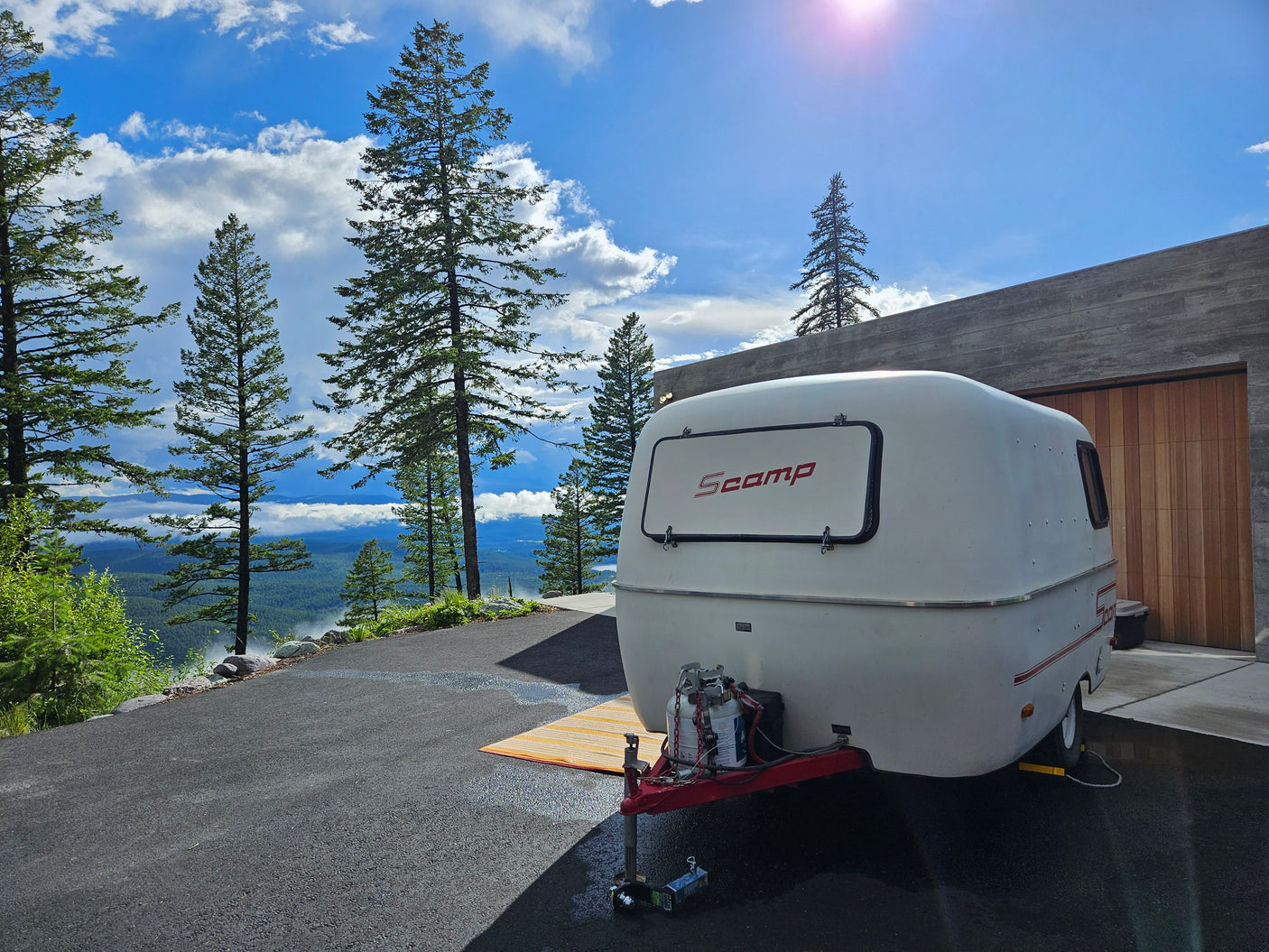 A white Scamp travel trailer, ideal for RV rental, overlooks a misty mountain valley from a modern driveway under a bright sky.