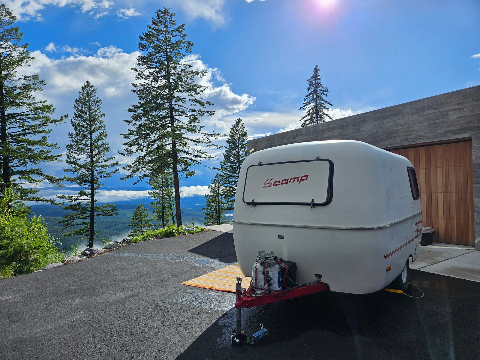 A white Scamp travel trailer, ideal for RV rental, overlooks a misty mountain valley from a modern driveway under a bright sky.