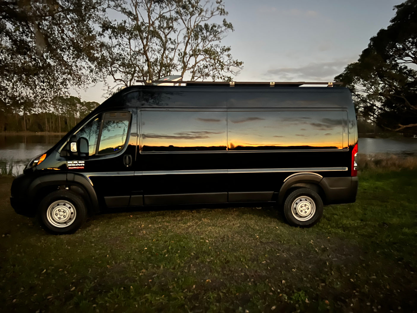 Black RV rental van reflects a colorful sunset over calm water. Ideal for scenic road trips and adventure travel.