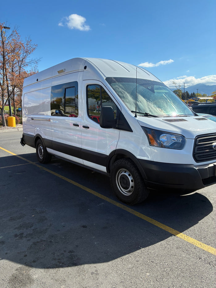 This white Ford Transit rv rental van, ready for adventures, is parked in a sunny lot. Reflected blue sky and fall trees.
