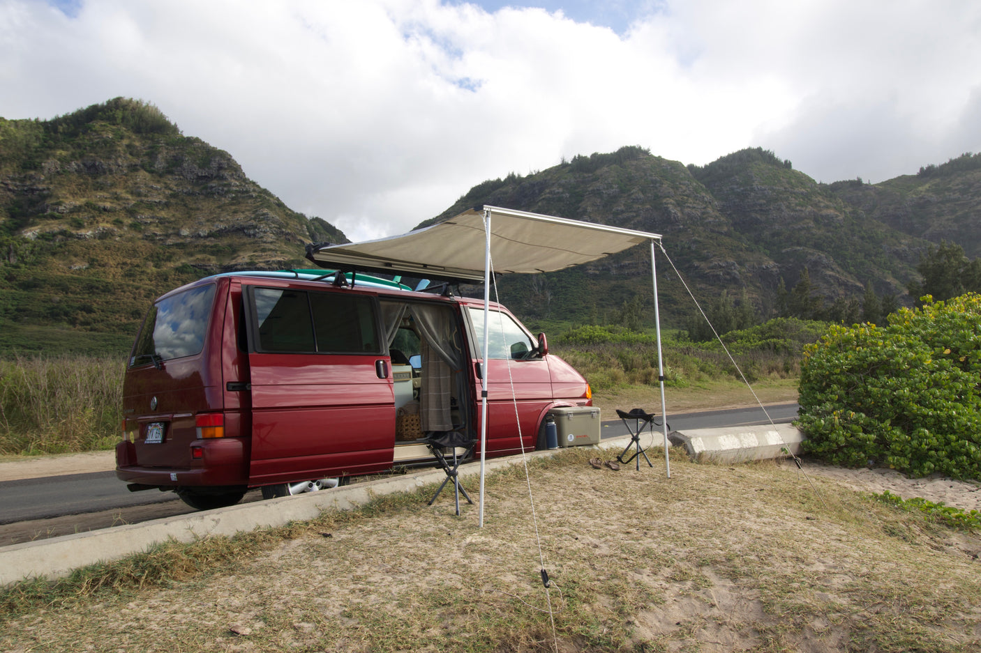 Red camper van with awning & surfboards on a scenic Hawaiian roadside. Perfect for your next RV rental adventure.